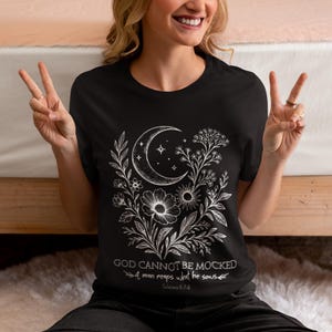 Puede incluir: Camiseta negra con un diseño floral blanco y una luna creciente. El texto "GOD CANNOT BE MOCKED. A man reaps what he sows" está impreso debajo. La camiseta está hecha de un material suave.