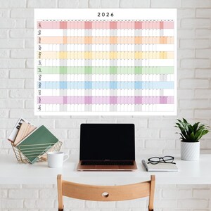 Peut inclure: Un calendrier mural 2026 avec un code couleur arc-en-ciel. Le calendrier est affiché sur un bureau blanc avec un ordinateur portable, des livres et une plante. Le calendrier a une mise en page en grille.