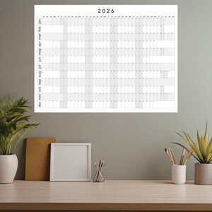 Peut inclure: Un grand calendrier mural blanc pour l'année 2026, avec une grille. Il est au-dessus d'un bureau en bois avec des éléments décoratifs, dont des plantes, un cadre photo et des articles de papeterie.