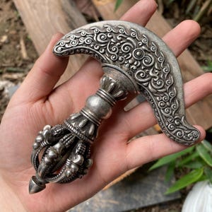 May include: A silver-toned ritual chopper with an ornate, curved blade featuring intricate swirling patterns. The handle is designed with a multi-pronged, cage-like structure and a pointed tip, showcasing detailed metalwork throughout the object.