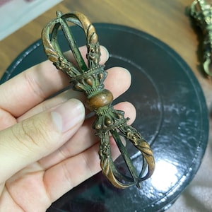 May include: A bronze-colored vajra, a ritual object with ornate detailing. The central section is bulbous, with intricate patterns and a loop at each end. The object is held in a hand, showcasing its size and craftsmanship.