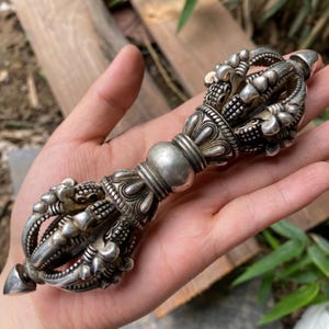 Puede incluir: Un objeto ritual vajra de metal color plata con una empuñadura esférica central flanqueada por dos extremos simétricos. Cada extremo consta de múltiples puntas curvas que se unen en un ápice, decoradas con patrones en relieve.