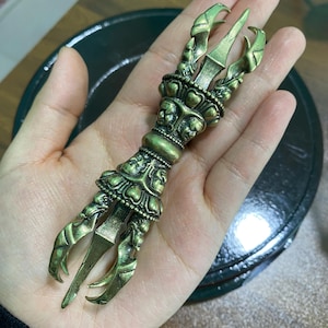 May include: A detailed bronze-colored claw-shaped object with intricate carvings. The claw has three main prongs and is decorated with floral and skull-like motifs. The object is held in a hand, showcasing its ornate design and craftsmanship.