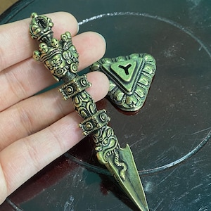 May include: An antique bronze-colored dagger with intricate carvings and a triangular base. The dagger features detailed patterns, including faces, skulls, and a serpent design. The triangular base has a Y-shaped design.