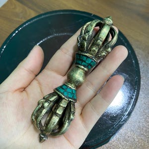 May include: A bronze-colored Vajra, a ritual object, with turquoise-colored accents. The Vajra features a central sphere and intricate, stylized hand-like prongs at each end. The object is held against a dark green background.