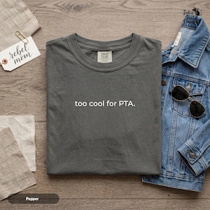 Op de afbeelding: Een antracietgrijs t-shirt met ronde hals en de witte tekst "too cool for PTA." op de borst gedrukt. Het shirt heeft een Comfort Colors merklabel aan de binnenkant van de halslijn. Het kledingstuk wordt getoond naast een blauw spijkerjasje en een zonnebril.
