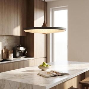 May include: A circular pendant light fixture with a dark gray outer rim and a white, pleated, translucent diffuser. The light is suspended by a thin, dark rod from the ceiling, casting a soft, warm glow downward over a kitchen island.