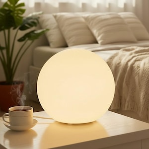 May include: A spherical table lamp with a smooth, matte white surface that emits a warm, soft glow. The lamp sits on a white surface next to a white ceramic cup and saucer containing a steaming hot beverage, creating a cosy and calm atmosphere.