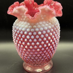 Puede incluir: Un jarrón de cristal rosa con clavos y un borde con volantes. El jarrón tiene un cuerpo redondo cubierto de pequeños puntos en relieve. El borde es de un rosa más oscuro y el cuerpo se desvanece de blanco a rosa. El jarrón se asienta sobre una base pequeña y redonda.