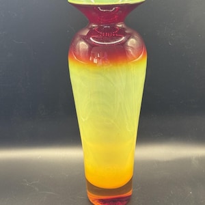 Puede incluir: Un jarrón de vidrio alto y colorido con una parte superior acampanada y una base redondeada. El jarrón hace la transición de una base roja oscura a un cuerpo amarillo-verdoso, con un degradado rojo y amarillo-naranja en el medio. El borde es verde.