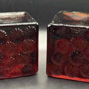 Puede incluir: Dos recipientes cuadrados de vidrio rojo rubí. Cada uno tiene una forma rectangular con una parte superior ligeramente ensanchada. Los lados están decorados con un patrón repetido de diseños florales en relieve. El vidrio tiene un acabado brillante.