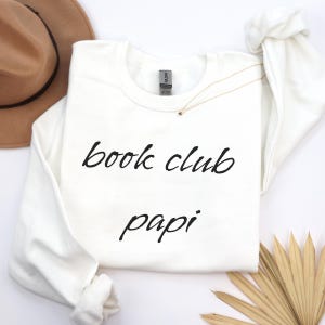 May include: A white sweatshirt with the words "book club papi" in black script. A gold necklace with a small star pendant is draped on the sweatshirt. A brown hat and dried plant are in the background.