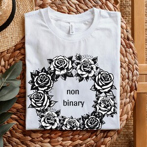 May include: White t-shirt featuring a black and white floral wreath design with the words "non binary" in the center. The wreath is composed of roses and leaves. The shirt is made of a soft material.