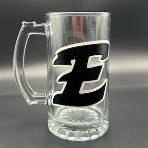 May include: Clear glass beer mug with a handle. The mug features a large black letter "E" with a white outline on the front. The mug has a thick base and a clear handle.