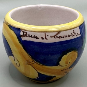 May include: A ceramic bowl with a yellow rim and a blue and yellow design. The bowl has a white interior and a label with the text "Duca di Camastra". The bowl is likely handmade and suitable for decorative use.