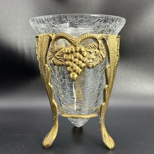 Puede incluir: Un florero de vidrio craquelado transparente dentro de un soporte de metal dorado. El marco de metal tiene tres patas y un diseño en relieve de un racimo de uvas con hojas en la parte frontal. El vidrio tiene una textura agrietada.