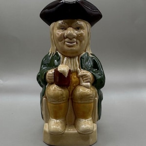 May include: A ceramic Toby jug figurine depicting a seated man in a tricorn hat, green coat, and yellow breeches, holding a mug of beer. The jug is tan, brown, and green.