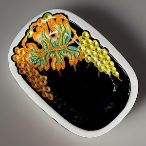 May include: An oval ceramic dish with a glossy black interior and a white outer rim. The interior features a hand-painted design of green and yellow grape clusters with large, detailed green and orange leaves against the dark black background.