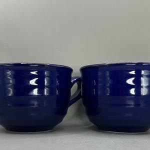 May include: Two cobalt blue ceramic mugs with handles. The mugs have a rounded shape and a textured exterior. The mugs are sitting on a white surface. The mugs are empty.