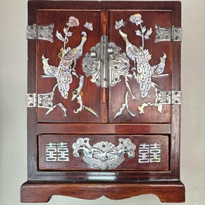 Può includere: Un portagioie in legno marrone scuro con due ante decorate con intarsi in madreperla raffiguranti pavoni e fiori, cerniere in metallo argentato e una chiusura centrale. Un cassetto sottostante presenta finiture argentate e simboli in madreperla.