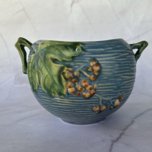 May include: A blue ceramic bowl with two handles and a textured surface. The bowl features a green leaf and brown berry design on the side. The bowl is a decorative piece.