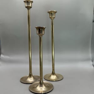 Puede incluir: Tres candelabros de latón de diferentes alturas sobre un fondo neutro. Cada candelabro tiene una base redonda, un tallo delgado y una parte superior en forma de copa para sostener una vela. Los candelabros son de un material metálico brillante.