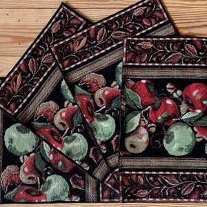 May include: A set of decorative placemats and a table runner featuring a fruit design. The placemats and runner have a black background with red apples, green apples, and green leaves. The borders are brown with a leaf pattern.