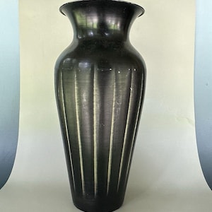 Peut inclure: Un grand vase en métal sombre avec une finition brossée et un bord évasé. Le corps du vase présente des cannelures verticales qui s'étendent de la base au col, créant un aspect texturé avec des reflets subtils le long des bords en relief.