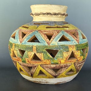 May include: A round ceramic vessel featuring a wide neck and a textured, cream-colored rim. The body is decorated with horizontal bands of cut-out triangles in shades of blue, green, yellow, and brown, creating a geometric, openwork pattern.