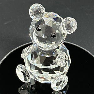 Puede incluir: Una figura de cristal transparente facetado con forma de oso. El oso tiene una cabeza redonda con dos orejas pequeñas, un cuerpo esférico y extremidades pequeñas. Presenta dos pequeñas cuentas negras opacas y lisas que sirven como ojos.