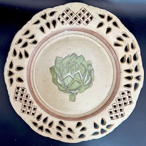 May include: A round ceramic plate with a cream-colored crackle glaze finish featuring a central green artichoke illustration. The wide rim has a decorative lattice pattern and cut-out teardrop shapes, accented by a thin brown circular border.