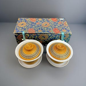 May include: Two lidded tea bowls with white ceramic saucers and woven bamboo covers featuring black geometric bands. The bowls are presented in a rectangular box with a blue, red, and gold floral pattern and light blue fabric ribbon closures.