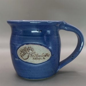 May include: A blue ceramic coffee mug with a curved handle. The mug features an oval emblem with the text "The Coffee Lab" and "Fairhope, AL" in a decorative font. The mug has a glossy finish.