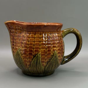 Könnte beinhalten: Ein Keramikkrug mit einem Maiskolben-Design. Der Krug ist hauptsächlich braun und gelb mit einem grünen Griff und Sockel. Der Körper des Kruges ist strukturiert, um Maiskörner nachzubilden.
