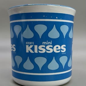May include: A cylindrical ceramic mug with a blue and white design. The mug features the words "Hershey's mini Kisses brand" in white lettering. The blue sections are decorated with light blue Hershey's Kiss-shaped graphics.