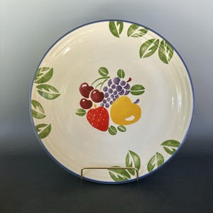 Puede incluir: Un plato de cerámica blanco con un borde azul y un diseño de frutas colorido. El plato presenta un grupo de cerezas, uvas, una fresa y una pera en el centro. Hojas verdes están pintadas alrededor del borde.