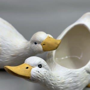 Peut inclure: Figurines de canards en céramique blanche avec des becs jaunes et des yeux bleus. L'un des canards a une cavité ouverte, peut-être pour contenir de petits objets. Les canards ont des détails de plumes texturés sur un fond neutre.