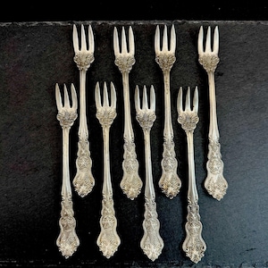 May include: A set of eight silver-toned metal cocktail forks arranged in two rows. Each fork features a three-pronged head and a handle decorated with an embossed floral and leaf pattern. The metal has a polished finish with intricate detailing.