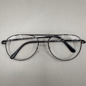 Puede incluir: Gafas de aviador de metal negro con lentes transparentes. Las gafas tienen un doble puente y el texto "54016 140 PD64 +/-2MM +2.00" está impreso en la barra superior. El texto "PL103 Paul GUN" está impreso en la lente derecha.