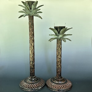 May include: Two metal candle holders shaped like palm trees with dark brown textured trunks and green leaf accents at the top. Each holder features a circular base with a cross-hatched pattern and a black cup at the center for holding a candle.