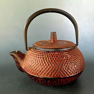 May include: A round cast iron teapot featuring a textured hobnail pattern across its surface. The teapot is finished in a gradient of deep red and black tones, includes a matching lid with a central knob, and has a curved metal handle.