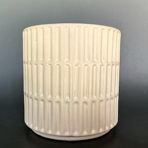 May include: A cylindrical ceramic planter in a creamy off-white color featuring a repeating vertical ribbed texture. The exterior surface is composed of raised, elongated bars arranged in a consistent pattern around the entire circumference.
