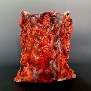 May include: A ceramic vase with a glossy, vibrant red and orange glaze. The surface features a highly textured, rippled, and folded appearance with dark brown and black accents integrated into the crevices of the undulating, organic clay form.
