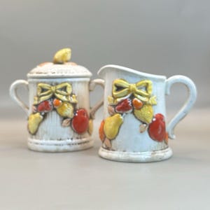 May include: A cream ceramic sugar bowl with lid and a creamer. Both are decorated with a yellow bow, red and yellow fruit, and green leaves. The sugar bowl has two handles.