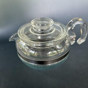 May include: A clear glass teapot with a matching lid and a curved handle. The teapot has a black band around the middle. The teapot is designed for brewing tea.