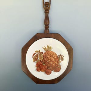 May include: A vintage trivet with a wooden frame and a ceramic tile featuring a pineapple and strawberries. The trivet has a dark brown wooden frame and a handle. The fruit illustration is in shades of brown, orange, and green.
