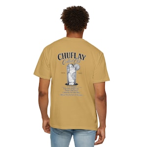 Op de afbeelding: Mosterdkleurig t-shirt met een cocktailafbeelding en de tekst "CHUFLAY Cocktail Club" op de rug. De afbeelding bevat een cocktailglas met limoenschijfjes en de tekst "ESTD 1920".