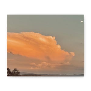 May include: A canvas print of a sunset scene. A large, fluffy orange cloud dominates the sky, contrasted against a pale blue background. The bottom of the image shows a silhouette of trees and mountains.