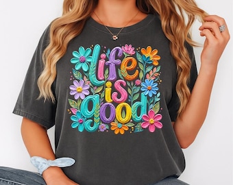 May include: Dark gray t-shirt with the phrase "life is good" in colorful, 3D lettering, surrounded by vibrant floral designs in pink, blue, orange, and purple. The shirt has short sleeves and a relaxed fit.