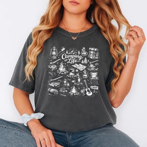 May include: Dark gray t-shirt with a white graphic of camping-related items, including a campfire, lantern, backpack, and guitar. The words "Camping Life" are in the center of the design. The shirt is a crew neck and has short sleeves.